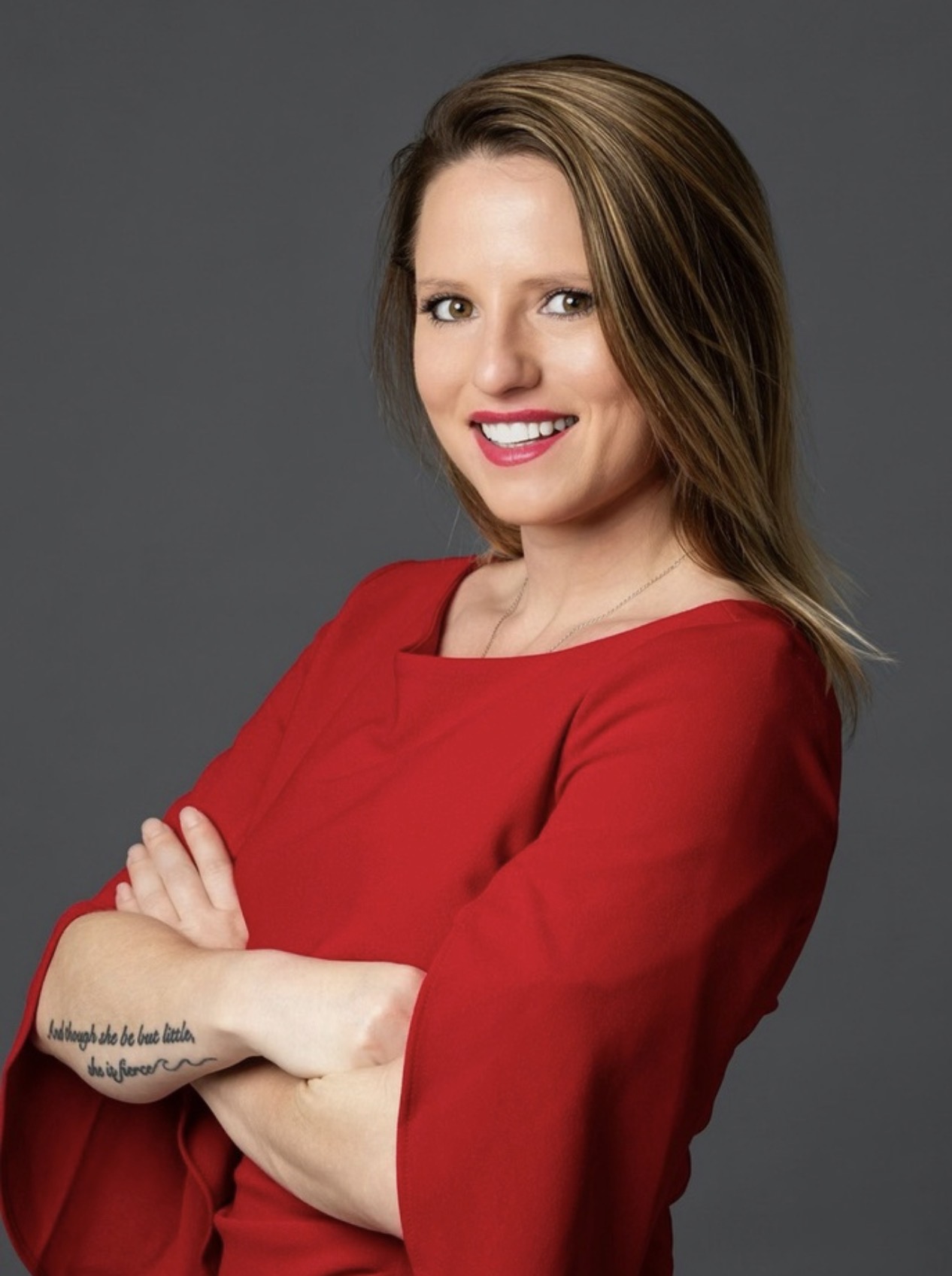 Confident woman with long highlighted hair wearing a red dress, arms crossed and smiling in a studio portrait against a gray background.